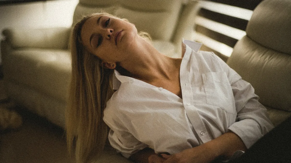 Femme en chemise blanche, la tête renversée sur un canapé, les yeux fermés, épuisée en fin de journée