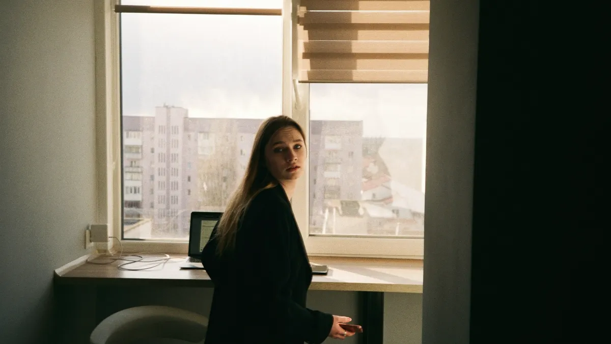 Femme en blazer noir dans son bureau, à demi dans l'ombre, jetant un regard inquiet par-dessus son épaule